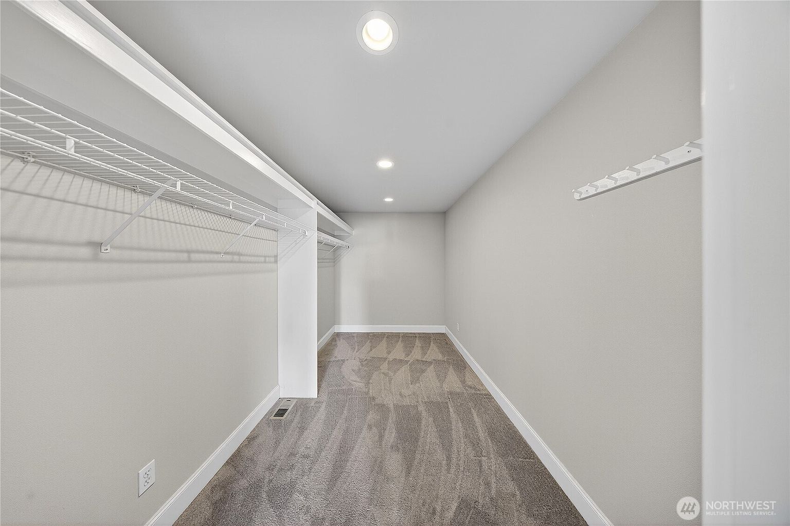This spacious walk-in closet features neutral gray carpeting and light gray walls, creating a clean and versatile storage area. The space is equipped with white wire shelving units along the left wall and a mounted coat rack on the right, providing ample organization options. Recessed lighting in the ceiling ensures the area is well-lit, offering a bright and functional walk-through perspective.