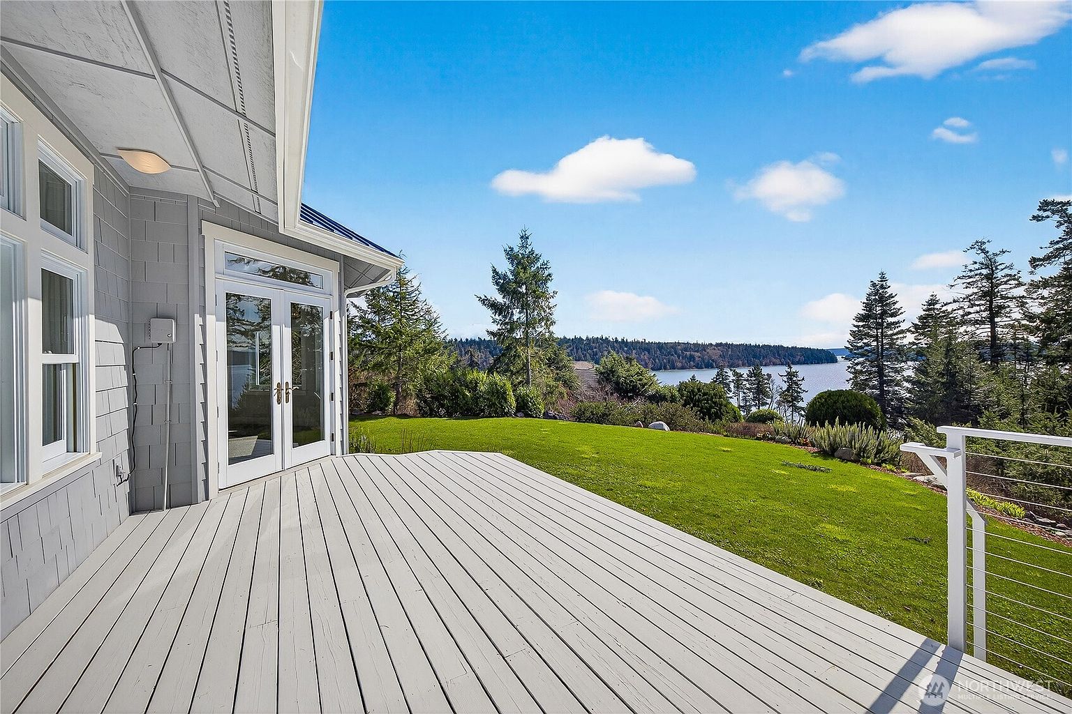 This image showcases a spacious, light-gray wooden deck attached to a modern home, featuring double glass doors that lead into the interior. The deck overlooks a lush green lawn that transitions into a scenic waterfront view with dense forest foliage in the background. The perspective is from the deck looking outward, capturing a serene and inviting outdoor living space perfect for relaxation or entertaining.