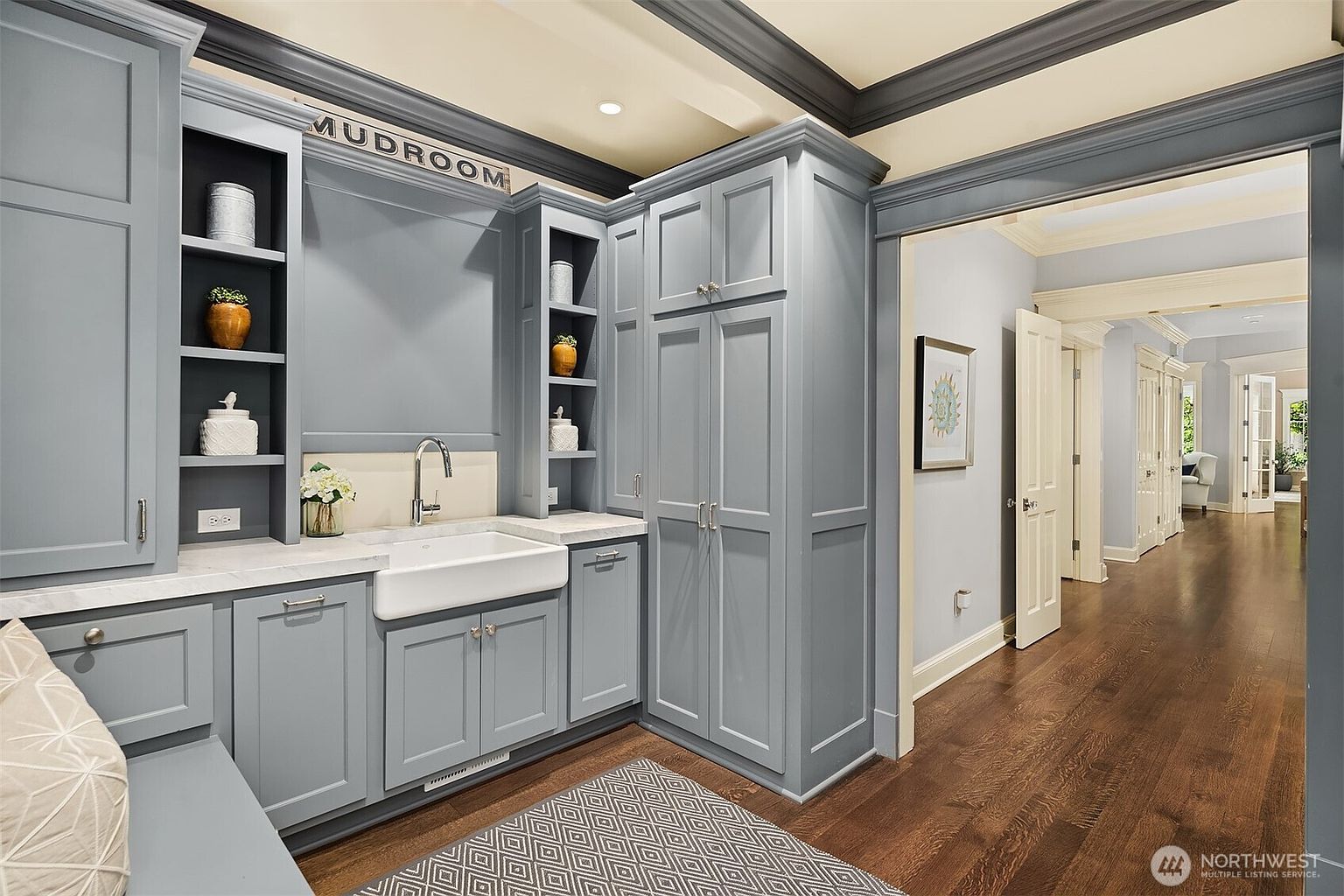 This elegant mudroom features custom blue-grey cabinetry with integrated shelving and a farmhouse-style sink set into a light-colored stone countertop. The space is accented by a patterned rug on dark hardwood floors and leads into a long, bright hallway. The overall aesthetic is sophisticated and functional, providing a clean transition area within the home.