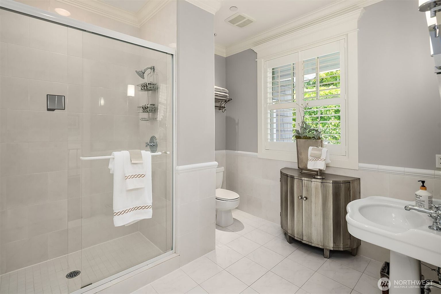 This bright and clean bathroom features a glass-enclosed walk-in shower with light-colored wall tiles and a built-in metal shelving unit. A pedestal sink sits to the right, complemented by a small, curved wooden vanity cabinet and a window with white shutters that allows natural light to fill the space. The room is finished with neutral-toned walls and light-colored floor tiles, creating a crisp and airy atmosphere.