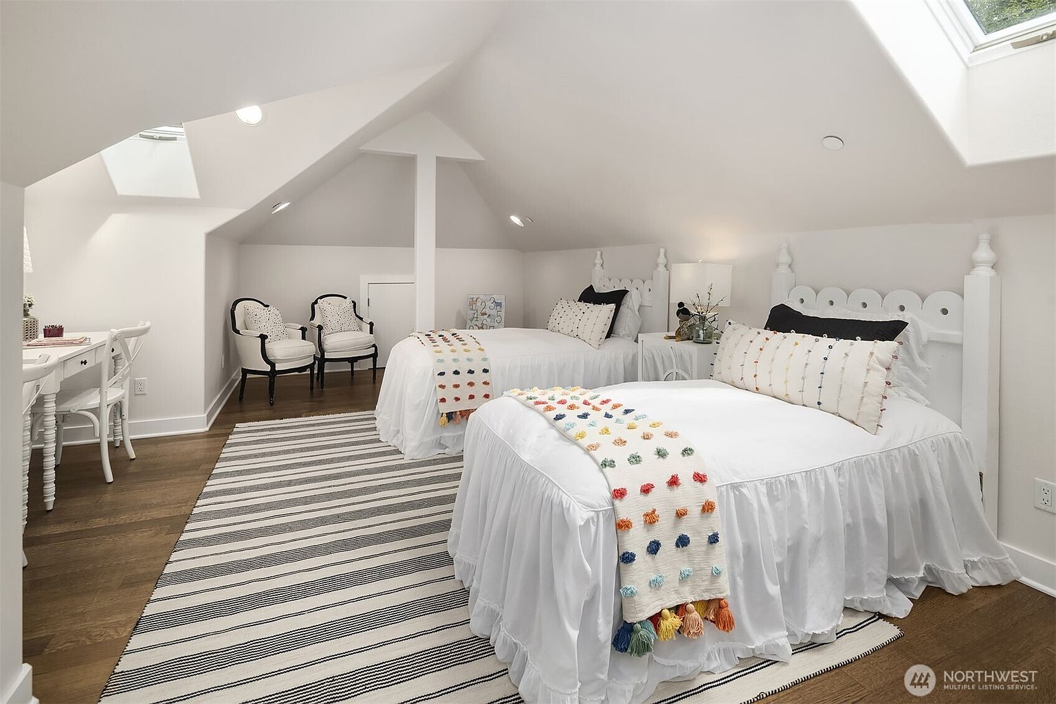This charming attic bedroom features two twin beds with white ruffled bedding and colorful pom-pom accents, set against a sloped ceiling with skylights. To the left, a small white desk and two elegant chairs create a cozy workspace, all anchored by a striped area rug on dark wood flooring. The room is bright and airy, utilizing the unique architectural angles of the attic to create a welcoming and functional living space.