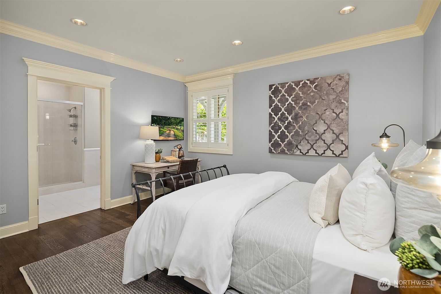 This well-appointed bedroom features light blue walls, dark wood flooring, and a metal-framed bed dressed in crisp white linens. A small desk area is tucked near the window, which is fitted with white plantation shutters, while a large, patterned wall art piece serves as a focal point above the bed. The room includes an open doorway leading to an attached bathroom with a glass-enclosed shower, creating a bright and cohesive living space.