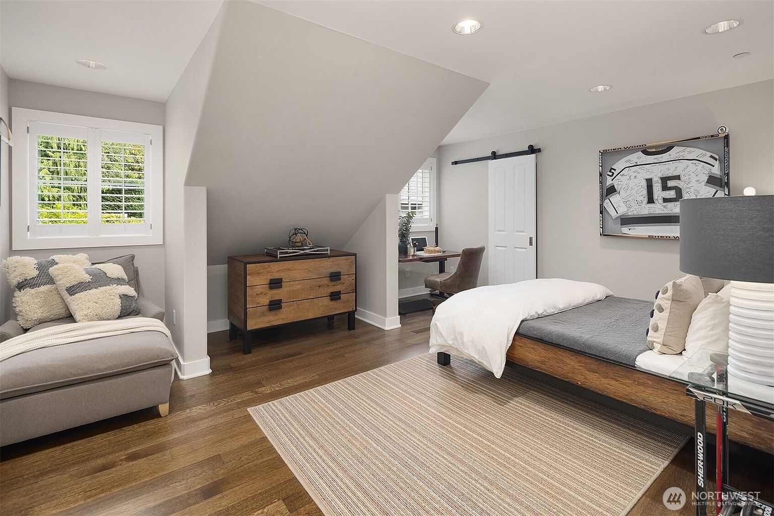This bedroom features a cozy, modern aesthetic with warm wood flooring and neutral gray walls. The room includes a bed with a wooden frame, a comfortable lounge chair with textured pillows, and a dedicated workspace tucked into an alcove under the sloped ceiling. A framed sports jersey on the wall adds a personalized touch to this well-lit, multi-functional living space.