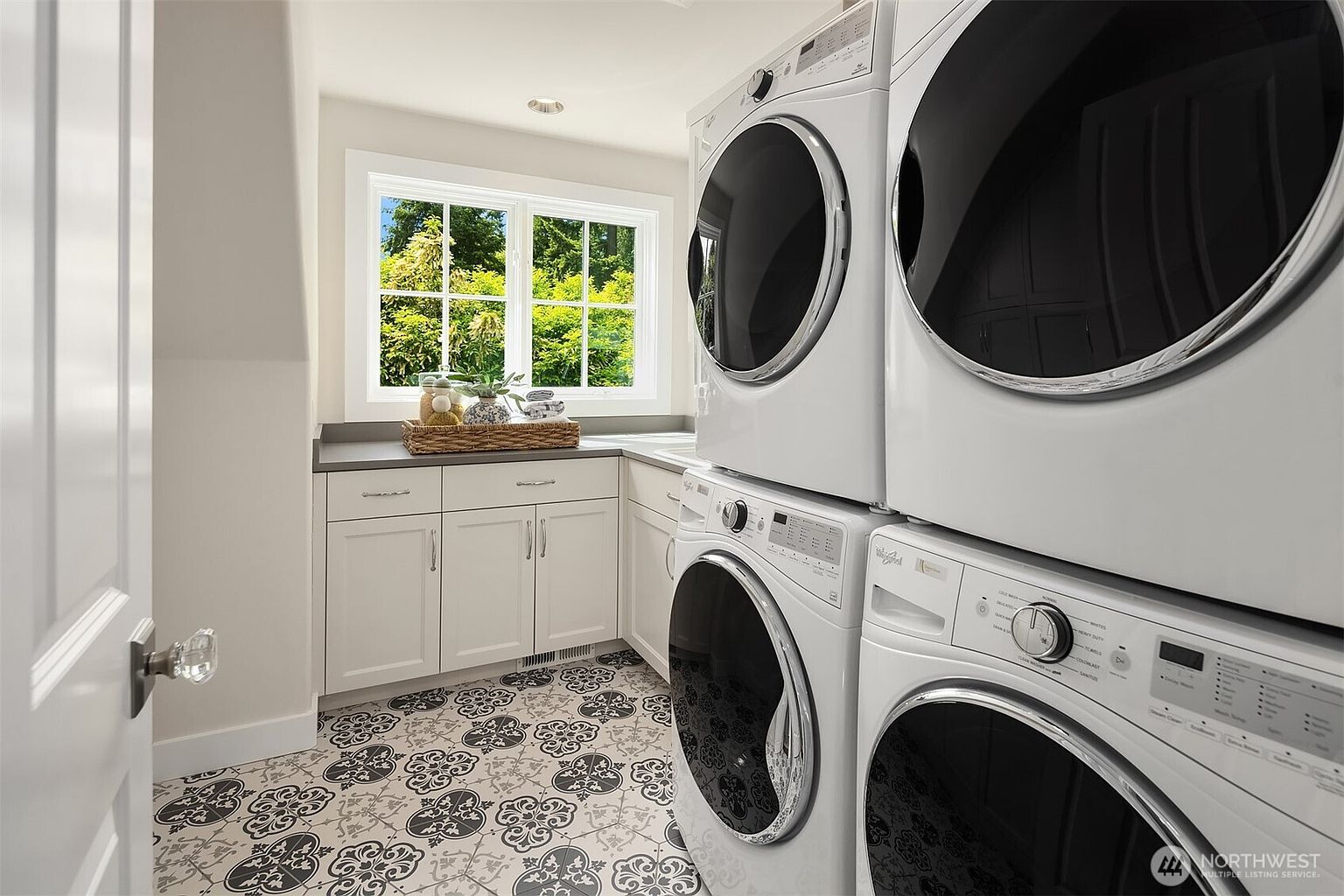This bright and functional laundry room features a stacked washer and dryer set positioned against a wall with a window overlooking greenery. The space includes white cabinetry providing ample storage, topped with a neutral countertop, and is finished with eye-catching patterned floor tiles. The perspective is from the doorway, offering a clear view of the efficient layout and clean, modern aesthetic.
