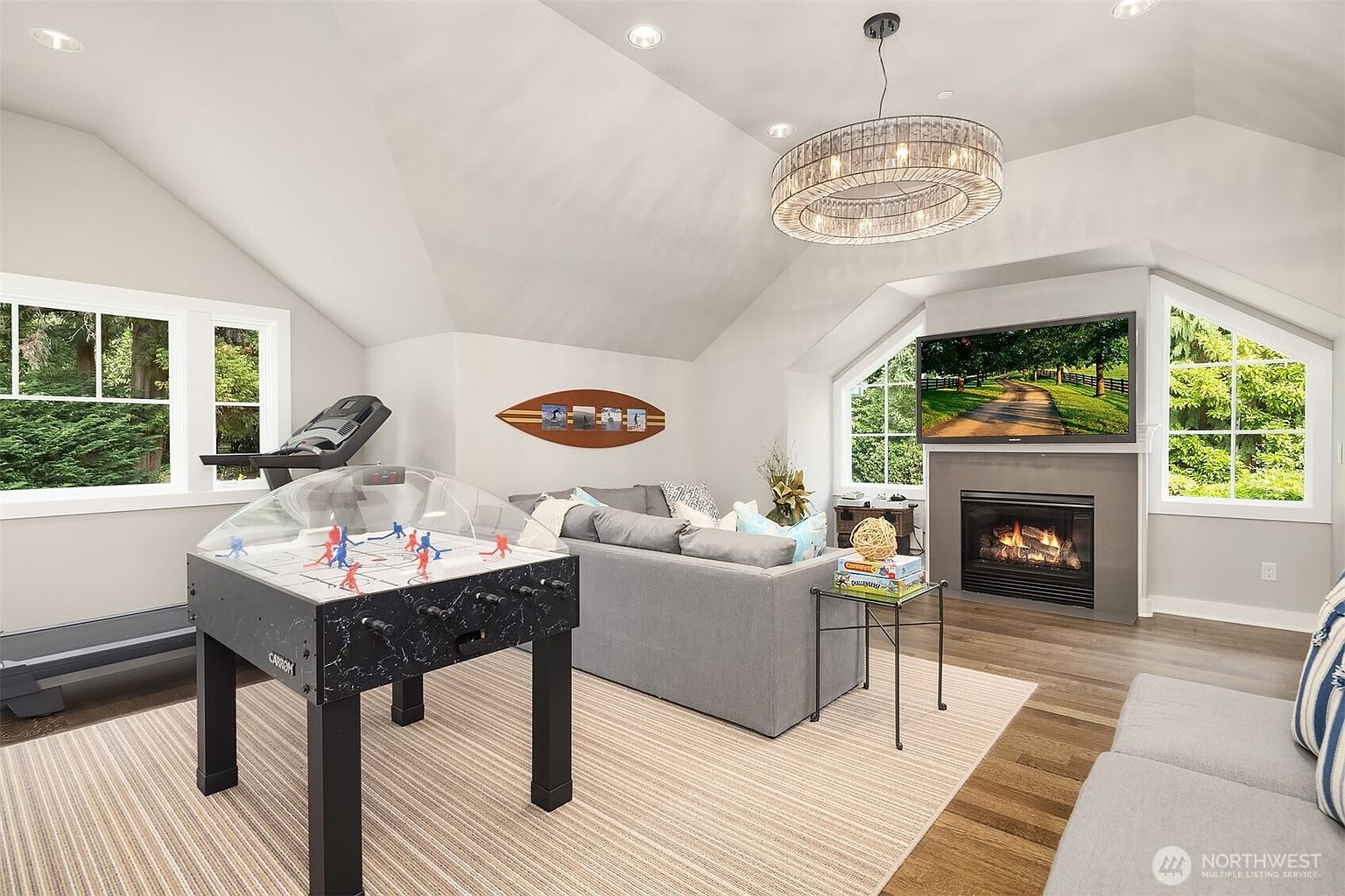 This spacious, multi-functional living area features a vaulted ceiling and a cozy seating arrangement centered around a fireplace with a dark, modern surround. The room is equipped for entertainment and recreation, showcasing a large wall-mounted television, a foosball table, and a treadmill tucked near the windows. Large windows on either side of the fireplace provide ample natural light and views of the surrounding greenery, creating a bright and inviting atmosphere.