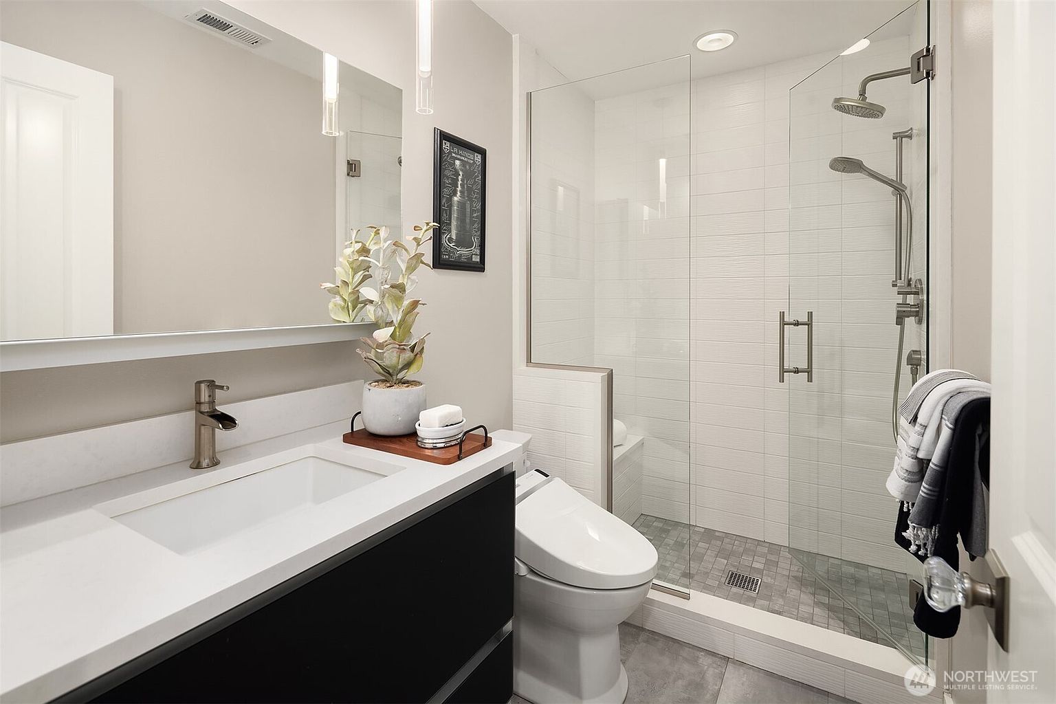 This modern bathroom features a sleek vanity with a dark base and a light-colored countertop, paired with a large mirror and contemporary pendant lighting. To the right, a glass-enclosed walk-in shower showcases white wall tiles and a gray tiled floor, complemented by a modern toilet with an integrated bidet seat. The space is accented with a small decorative plant and a framed piece of art, creating a clean and polished aesthetic.