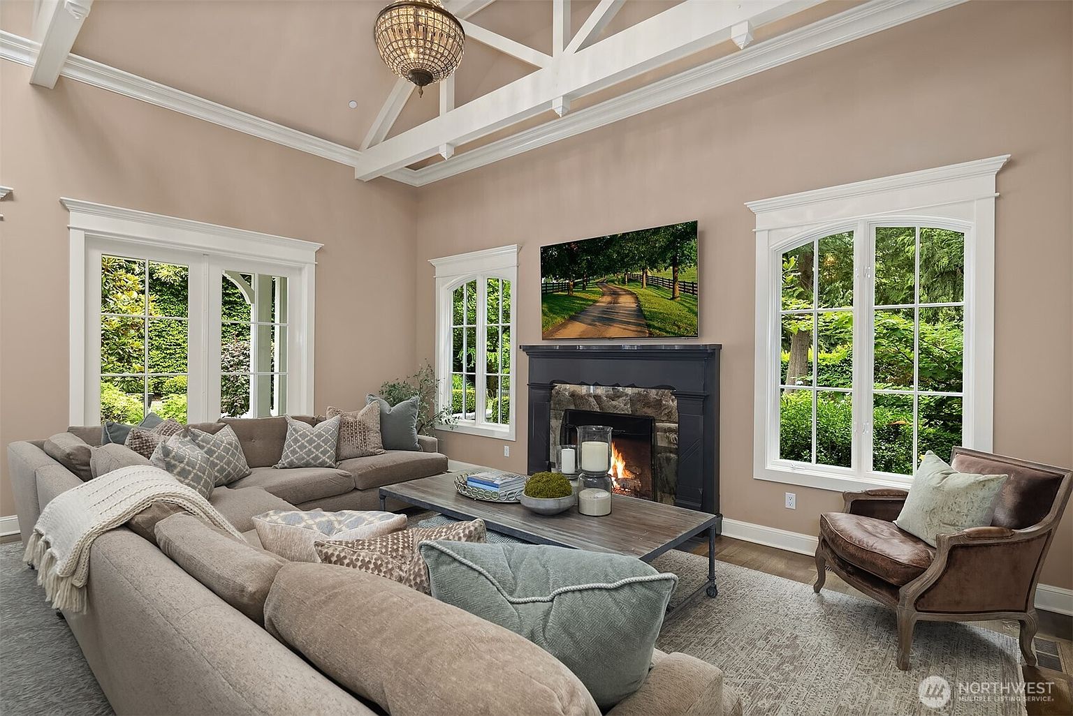 This inviting living room features a vaulted ceiling with exposed white beams and a central chandelier, creating an airy and sophisticated atmosphere. A large, plush sectional sofa faces a dark-painted fireplace surround with a stone hearth, while tall, arched windows provide abundant natural light and views of the lush greenery outside. The space is anchored by a textured area rug and a rustic wood coffee table, offering a comfortable and stylish setting for relaxation.
