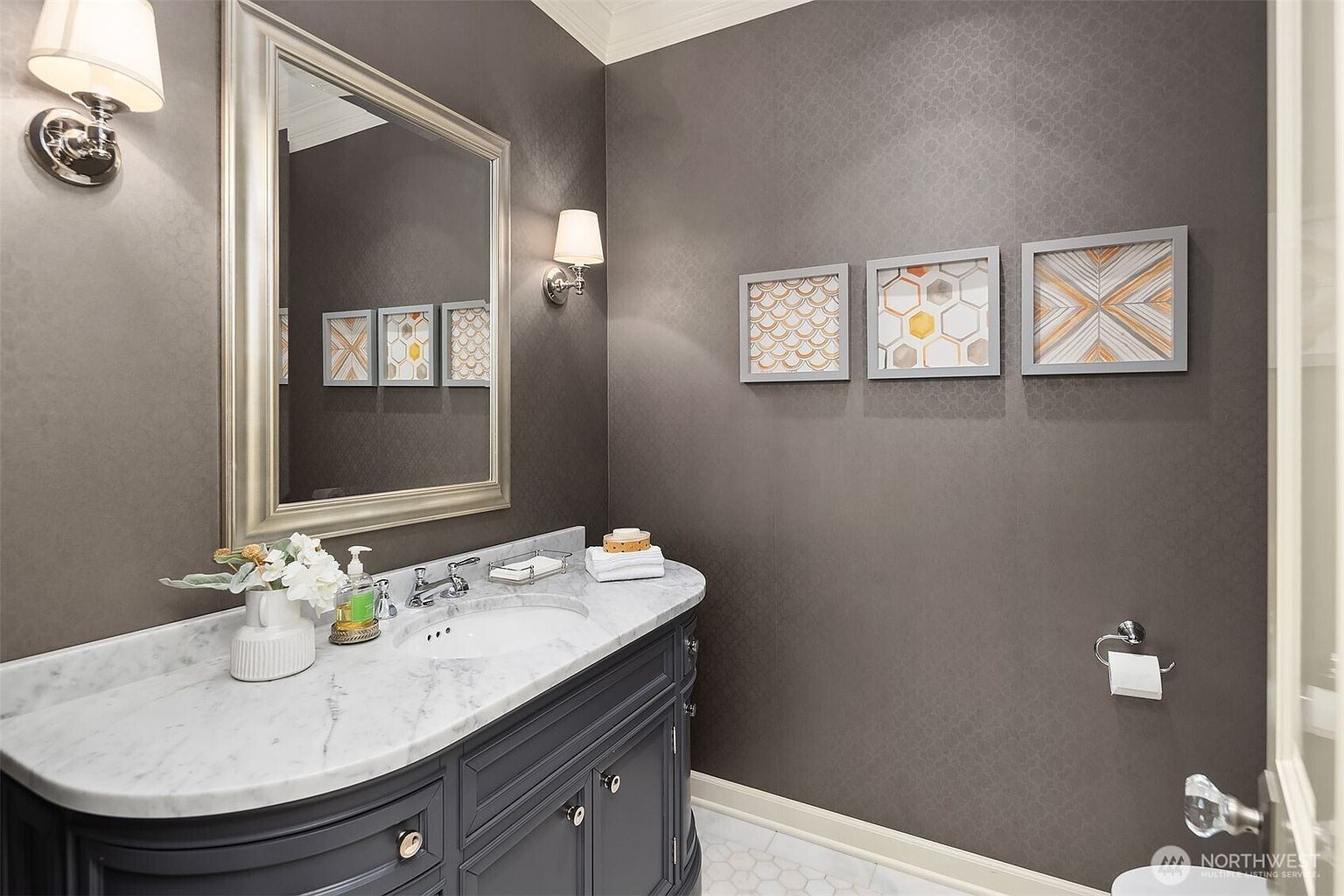This elegant powder room features dark, textured wallpaper and a vanity with a stone countertop and an undermount sink. A large, silver-framed mirror is flanked by two wall-mounted sconces, while three framed geometric art pieces add a decorative touch to the wall. The space is finished with a light-colored tile floor, creating a sophisticated and polished aesthetic.