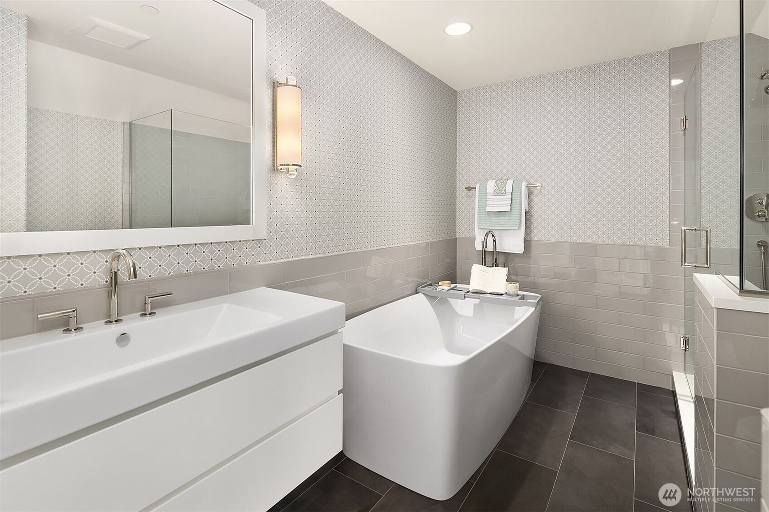 This modern primary bathroom features a sleek, white floating vanity paired with a freestanding soaking tub. The walls are finished with a combination of gray subway tile and a decorative patterned tile, while the floor is laid with dark, large-format tiles. A glass-enclosed shower is visible to the right, and a large mirror above the vanity enhances the bright, contemporary aesthetic of the space.