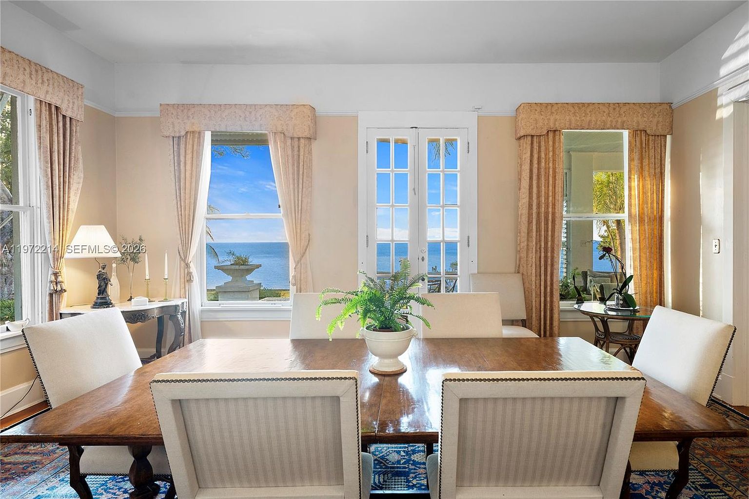 This elegant dining room features a large wooden table centered in the space, surrounded by upholstered chairs with nailhead trim. The room is framed by tall windows and a central set of French doors that provide a clear, expansive view of the ocean. A patterned area rug anchors the space, while soft, neutral wall tones and classic window treatments create a sophisticated and serene atmosphere.