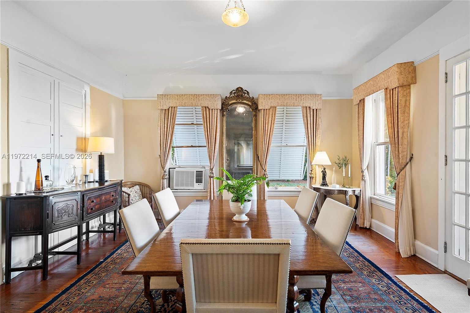 This formal dining room features a long wooden table surrounded by six upholstered chairs, set atop a large, intricately patterned area rug. The space is anchored by a tall, ornate gold-framed mirror positioned between two windows dressed in elegant curtains, while a dark wood sideboard provides additional storage and display space. The room is illuminated by a central pendant light and two table lamps, creating a warm and traditional atmosphere.
