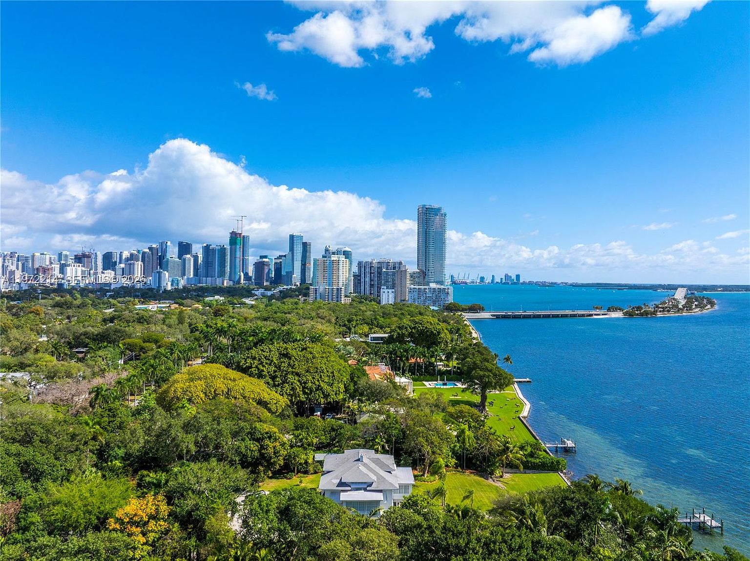 This high-angle aerial view captures a sprawling waterfront estate nestled within a lush, tree-filled landscape. The property features a large residence with a light-colored roof, expansive green lawns, and direct access to the bay, with a prominent city skyline visible in the distance. The perspective highlights the prime coastal location, emphasizing the blend of private greenery and urban proximity.