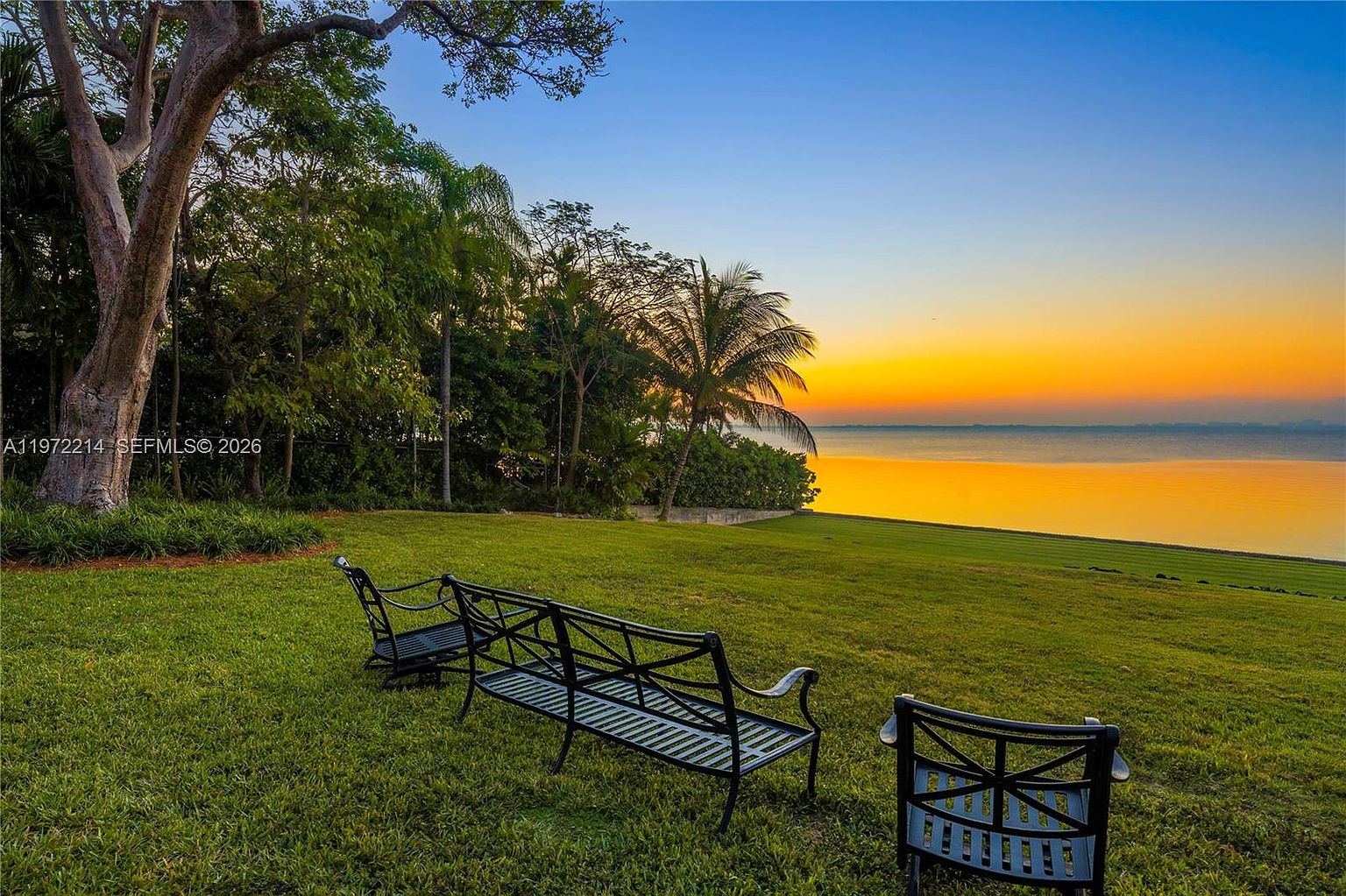 This serene outdoor space features a lush, expansive green lawn overlooking a calm body of water at sunset. Three black metal benches are arranged on the grass, providing a peaceful vantage point to enjoy the vibrant orange and blue sky. Mature trees and tropical palms frame the scene, creating a private and tranquil atmosphere.