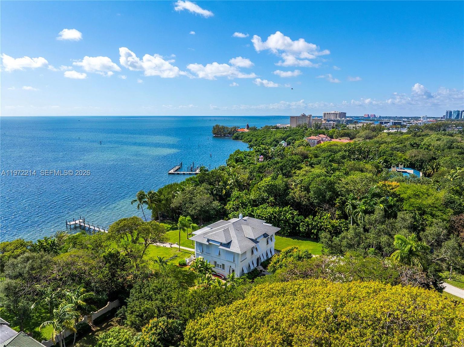 This high-angle aerial view captures a large, multi-story residence nestled within a lush, tree-filled coastal landscape. The property features a private dock extending into the expansive blue water, with a sprawling lawn surrounding the home. The scene offers a serene, panoramic perspective of the waterfront estate and the distant shoreline under a bright, clear sky.