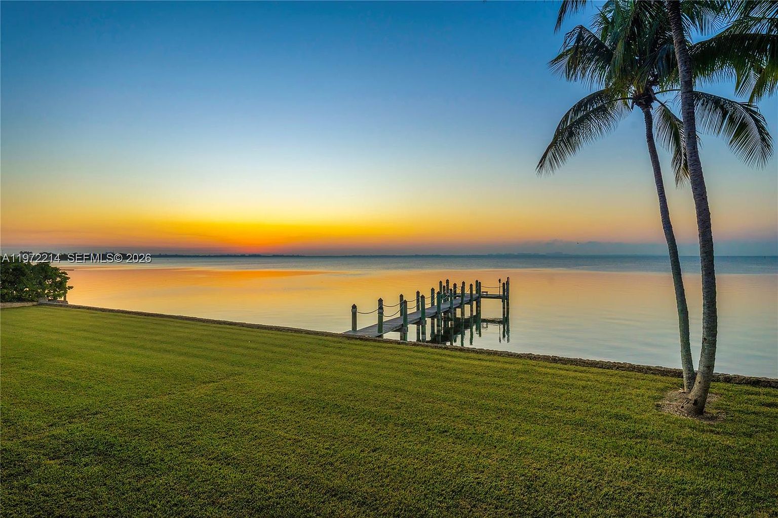 This serene waterfront view captures a lush, expansive green lawn leading directly to the calm, reflective waters of a bay at sunset. A wooden pier extends into the water, providing a focal point against the vibrant orange and blue gradient of the sky. Two tall palm trees frame the right side of the composition, enhancing the tropical, tranquil atmosphere of the property.