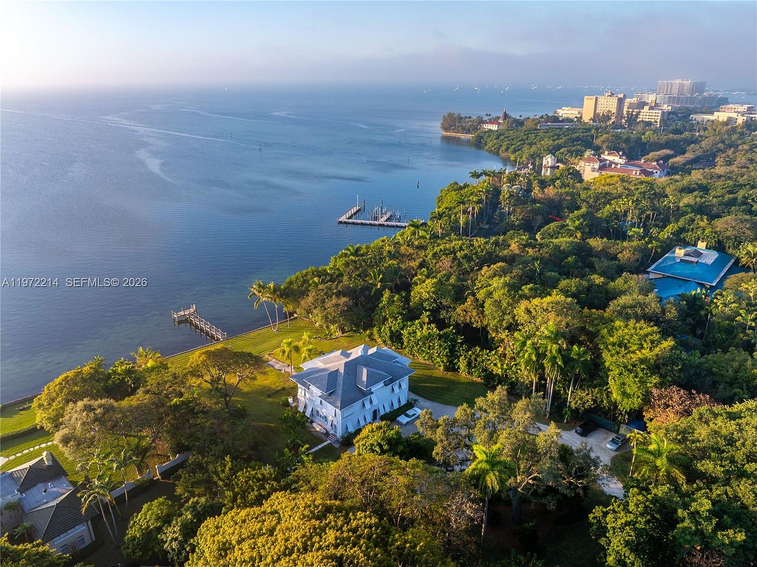 This high-angle aerial view captures a sprawling waterfront estate nestled within a lush, dense canopy of mature trees. The property features a classic multi-story residence with a light-colored exterior, a private wooden dock extending into the calm bay, and expansive green lawns. The perspective highlights the serene coastal setting, showcasing the seamless integration of the home into its natural, wooded surroundings.