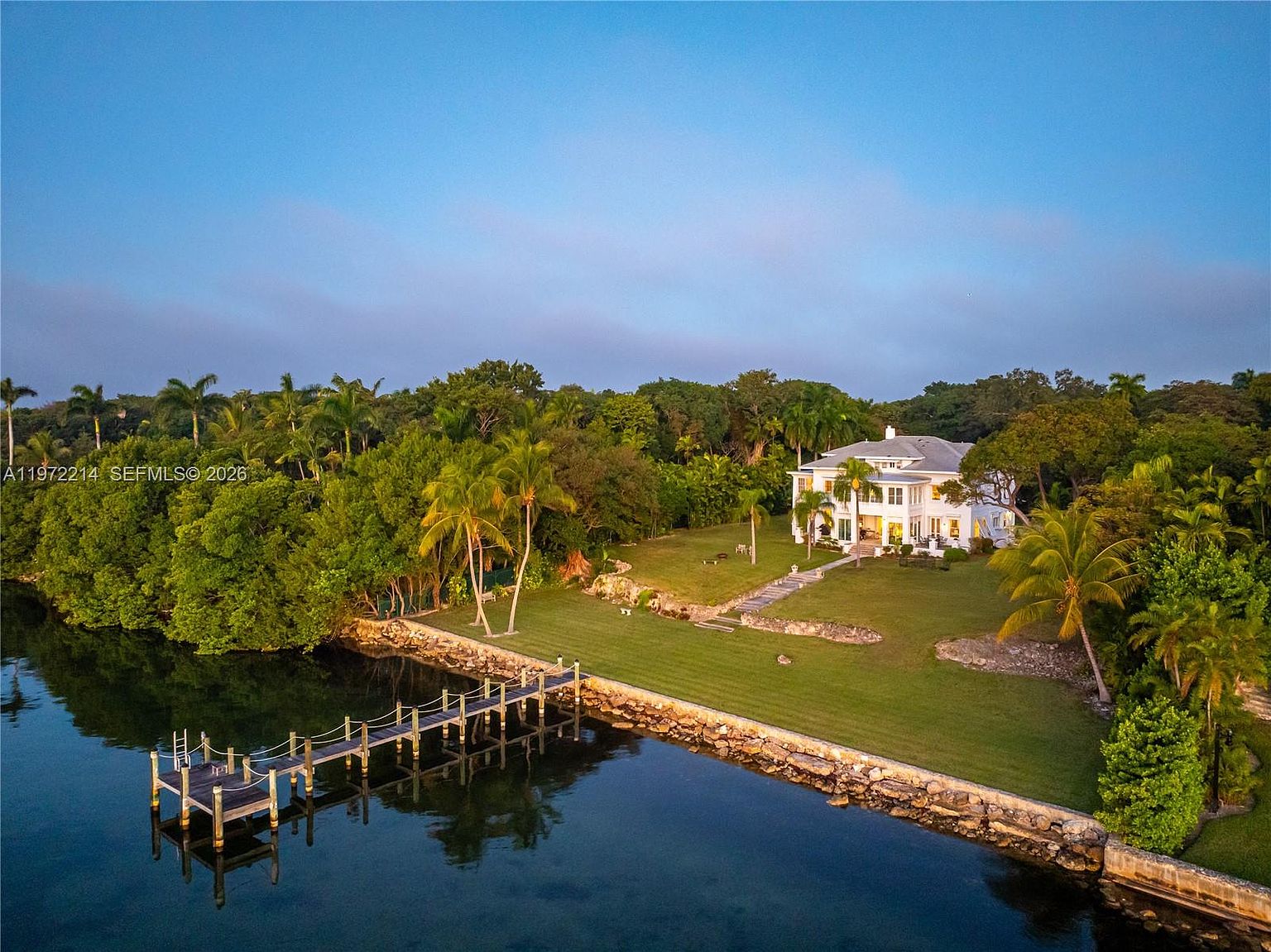 This aerial perspective captures a stately white multi-story residence situated on a lush, expansive waterfront lot. The property features a manicured lawn, mature tropical landscaping, and a private wooden dock extending into the calm water. The scene is bathed in the warm, golden light of late afternoon, highlighting the home's classic architectural design and its serene, secluded setting.
