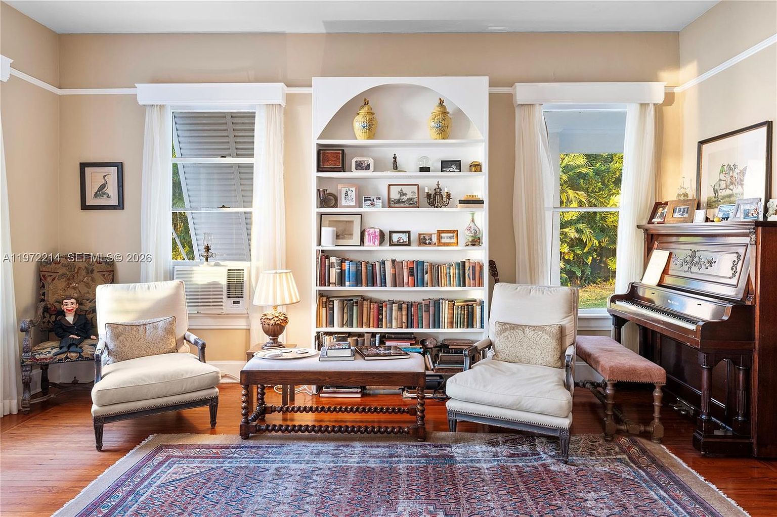 This cozy living room features a traditional aesthetic with hardwood floors and a large, central built-in bookshelf filled with books and decorative items. Two cream-colored armchairs flank a low, wooden coffee table, while an upright piano sits to the right, creating a warm and inviting atmosphere. The room is framed by two windows with light curtains, allowing natural light to illuminate the space and the patterned area rug.