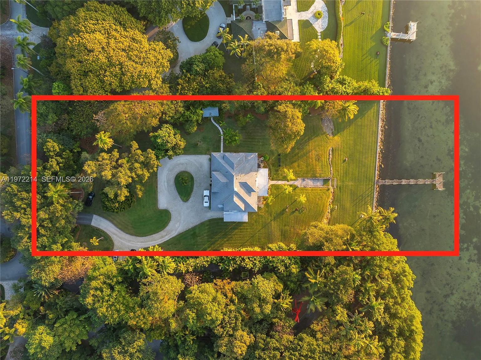 This high-angle aerial view captures a sprawling waterfront estate surrounded by lush, mature trees and expansive green lawns. The property features a long, winding driveway leading to a central residence, a private wooden dock extending into the water, and a serene, secluded atmosphere. The perspective highlights the generous lot size and the seamless integration of the home into its natural, verdant landscape.