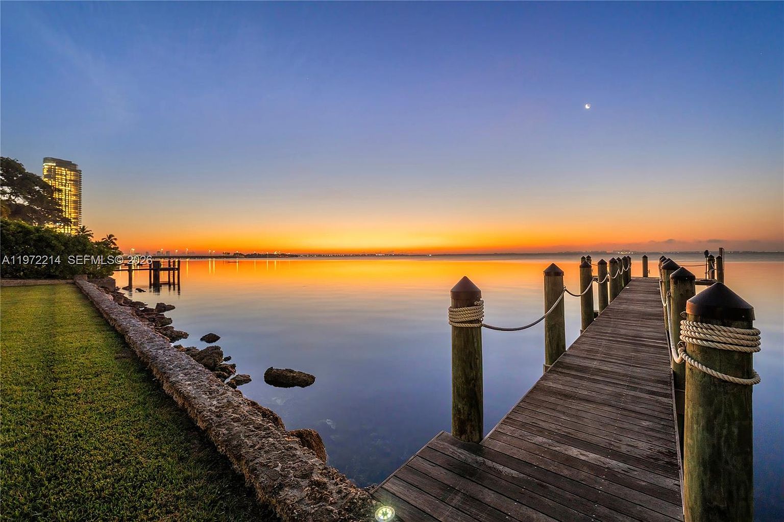 This serene waterfront view captures a wooden pier extending into a calm bay during a vibrant sunset. The foreground features a grassy lawn and a textured stone retaining wall, while the horizon is illuminated by warm orange and purple hues. A distant high-rise building is visible on the left, adding a touch of urban contrast to the tranquil natural setting.