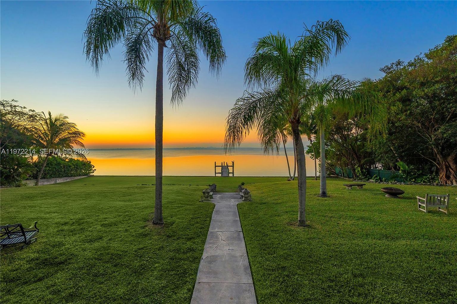 This expansive backyard features a lush green lawn leading directly to a serene waterfront view at sunset. A concrete pathway cuts through the center of the grass, flanked by mature palm trees and leading toward a small wooden dock at the water's edge. The scene is framed by dense foliage on the sides, creating a private and tranquil atmosphere with a vibrant orange and yellow sky.