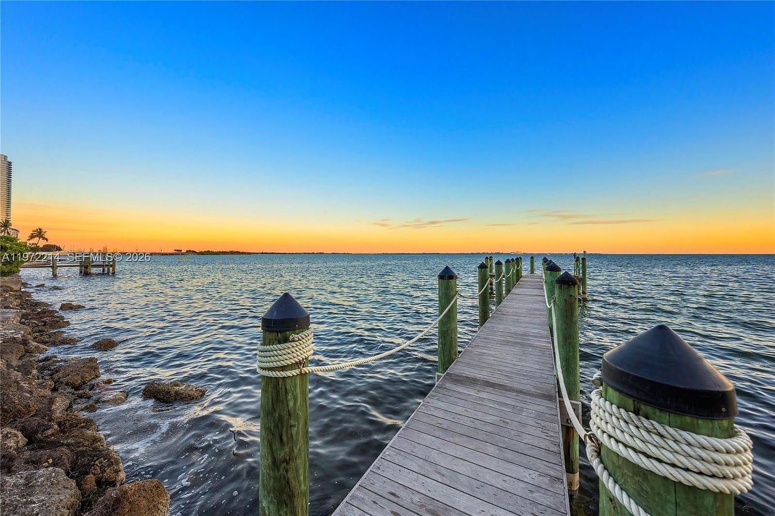 A wooden pier extends into calm, open water, leading the eye toward a vibrant sunset sky transitioning from orange to deep blue. The foreground features sturdy wooden pilings wrapped in thick rope, set against a rocky shoreline. This serene waterfront scene captures a peaceful coastal atmosphere, ideal for relaxation or boating access.