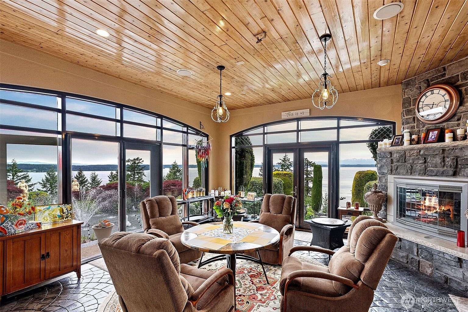 This sunroom features a warm, light wood-paneled ceiling and expansive windows that provide panoramic views of the water and garden. A stone-clad chimney breast anchors the room, housing a fireplace that adds a cozy element to the space. The room is furnished with comfortable seating arranged around a central table, creating an inviting atmosphere for relaxation while enjoying the scenery.