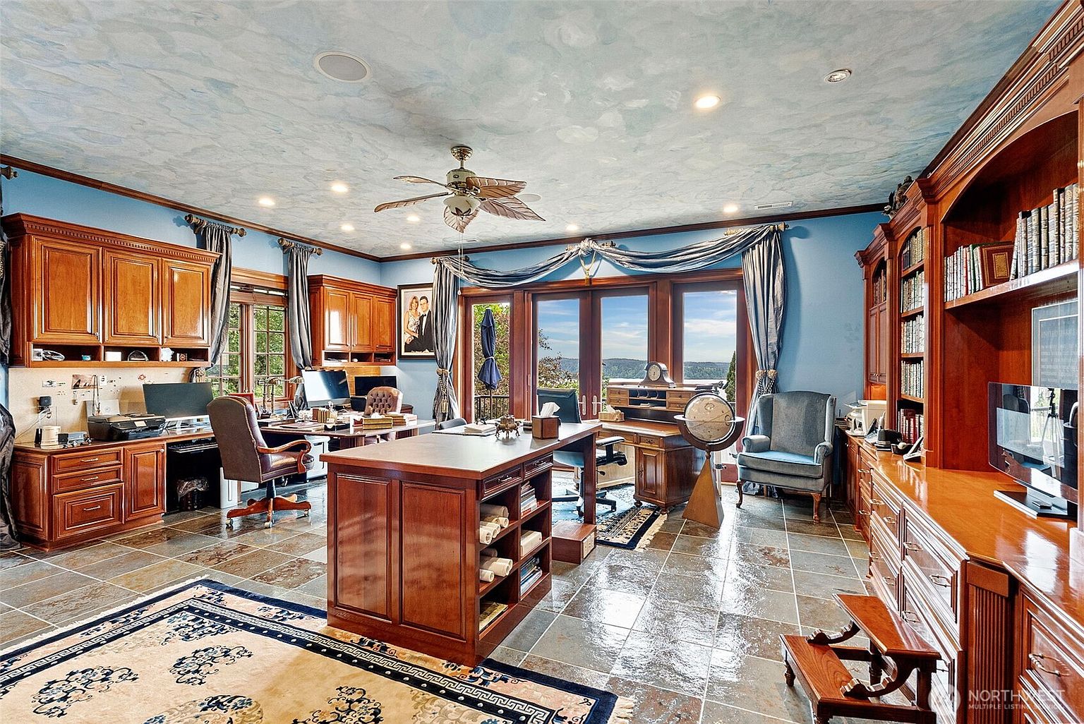 This spacious home office features a large central wooden desk, built-in cabinetry, and a decorative painted ceiling with a ceiling fan. The room is anchored by a patterned area rug over stone tile flooring and includes a dedicated workspace with a computer and a separate desk area near the windows. Large glass doors provide a view of the outdoors, creating a bright and professional atmosphere.