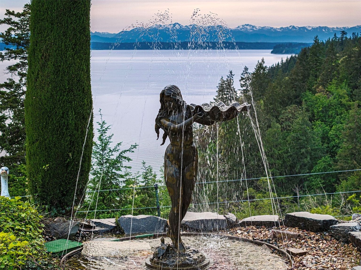 This serene garden scene features a bronze-toned statue of a woman holding a shell, serving as a central fountain element. The fountain is set against a breathtaking backdrop of a calm body of water and distant, snow-capped mountain peaks. Lush greenery, including a tall, slender cypress tree and dense forest, frames the view, creating a tranquil and picturesque outdoor setting.