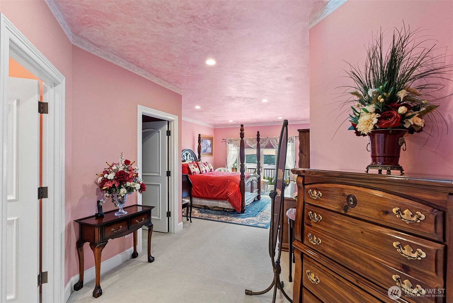 This primary bedroom suite features a distinctive pink color palette on the walls and ceiling, complemented by a light-colored carpet. The room is furnished with a dark wood four-poster bed, a matching wooden dresser, and a small console table topped with a floral arrangement. A doorway leads into the bedroom area, which includes a view of a balcony through large windows, creating a bright and traditional atmosphere.