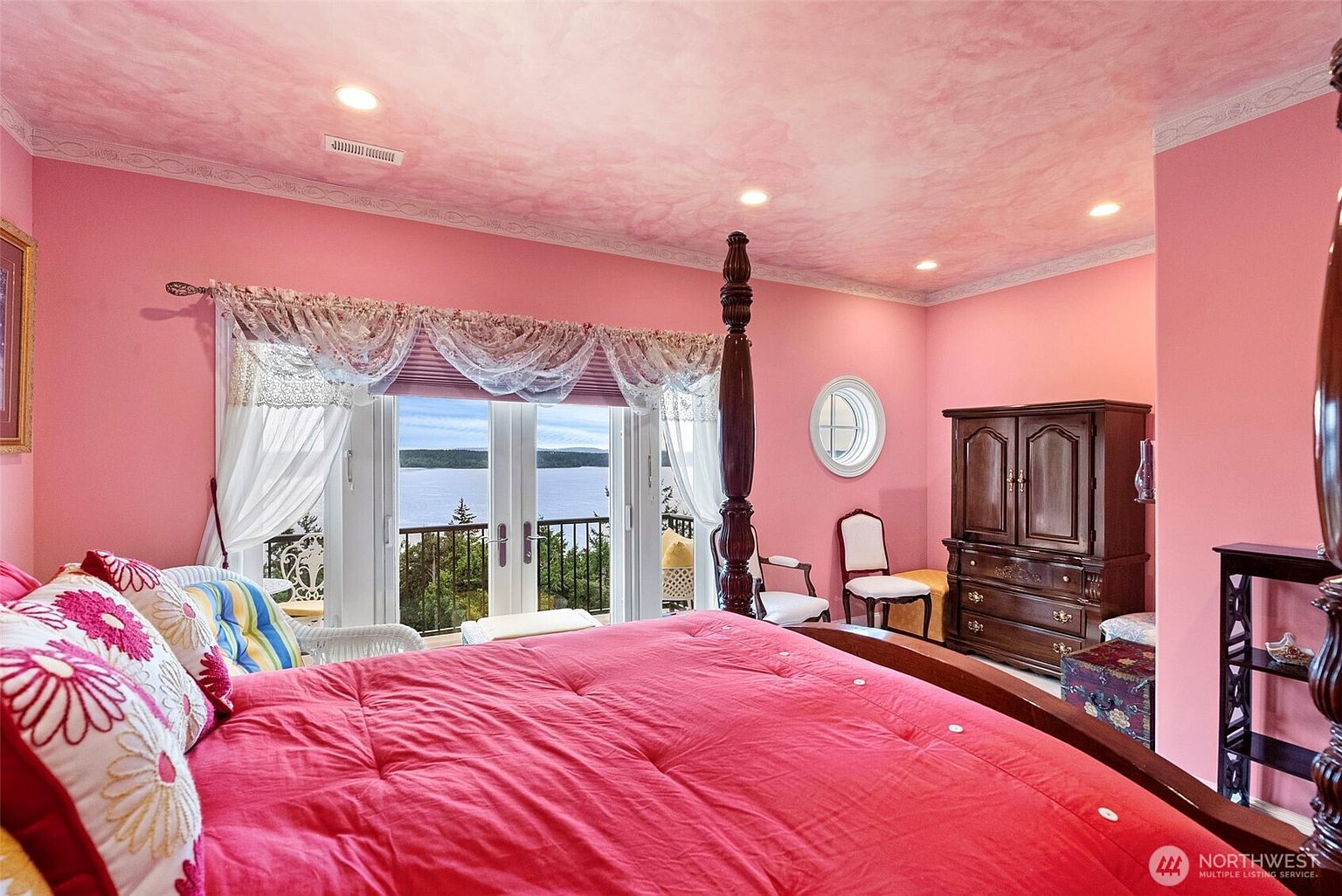 This primary bedroom features a bold, monochromatic pink color scheme with a textured ceiling and a large four-poster bed as the central focal point. A set of glass doors leads to a balcony, providing a scenic view of the water and surrounding trees. The room is furnished with a dark wood armoire and classic chairs, creating a traditional and vibrant atmosphere.