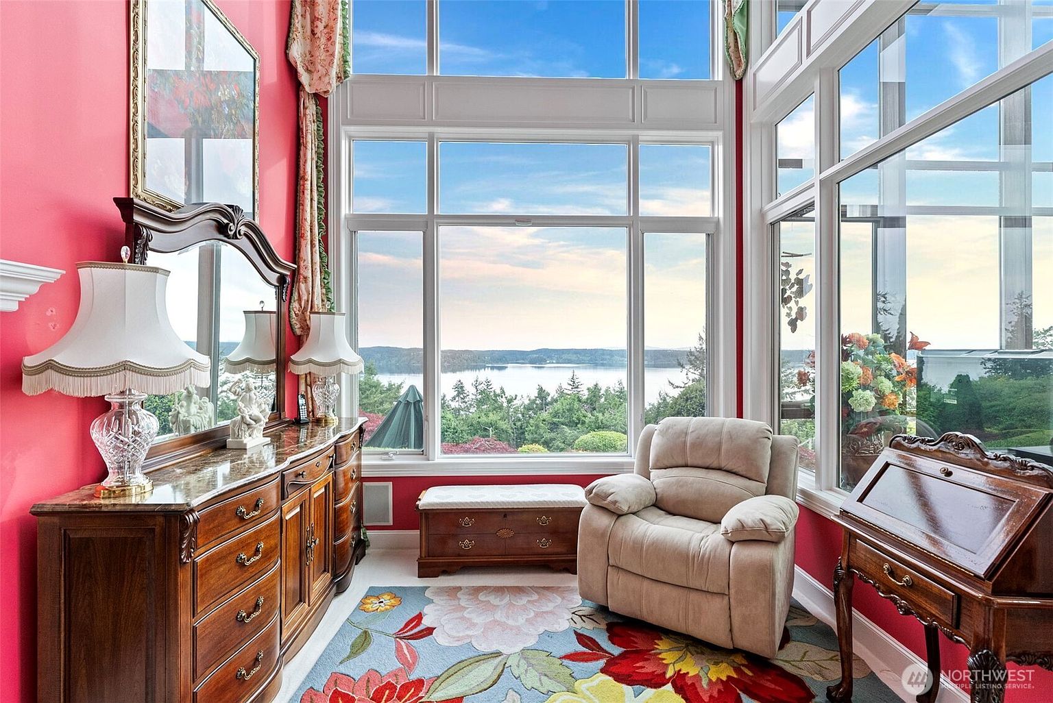 This sun-drenched room features vibrant red walls and a large, colorful floral area rug that anchors the space. A comfortable beige recliner sits beside a wooden writing desk, while a long dresser with a mirror provides ample storage and a classic aesthetic. The room is defined by expansive windows that offer a panoramic view of the surrounding trees and water, creating a serene and bright atmosphere.