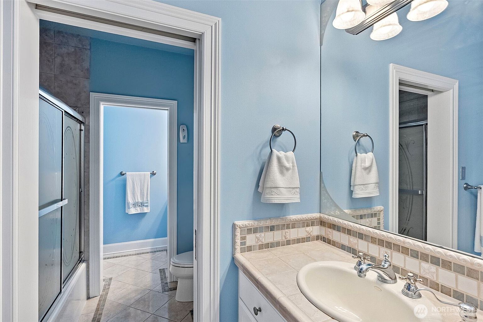 This bathroom features light blue walls and a vanity with a neutral-toned countertop and an integrated sink. A large mirror reflects the space, including a doorway leading to a separate area with a toilet and a shower-tub combination. The room is accented with decorative tile work along the backsplash and a classic light fixture above the mirror.