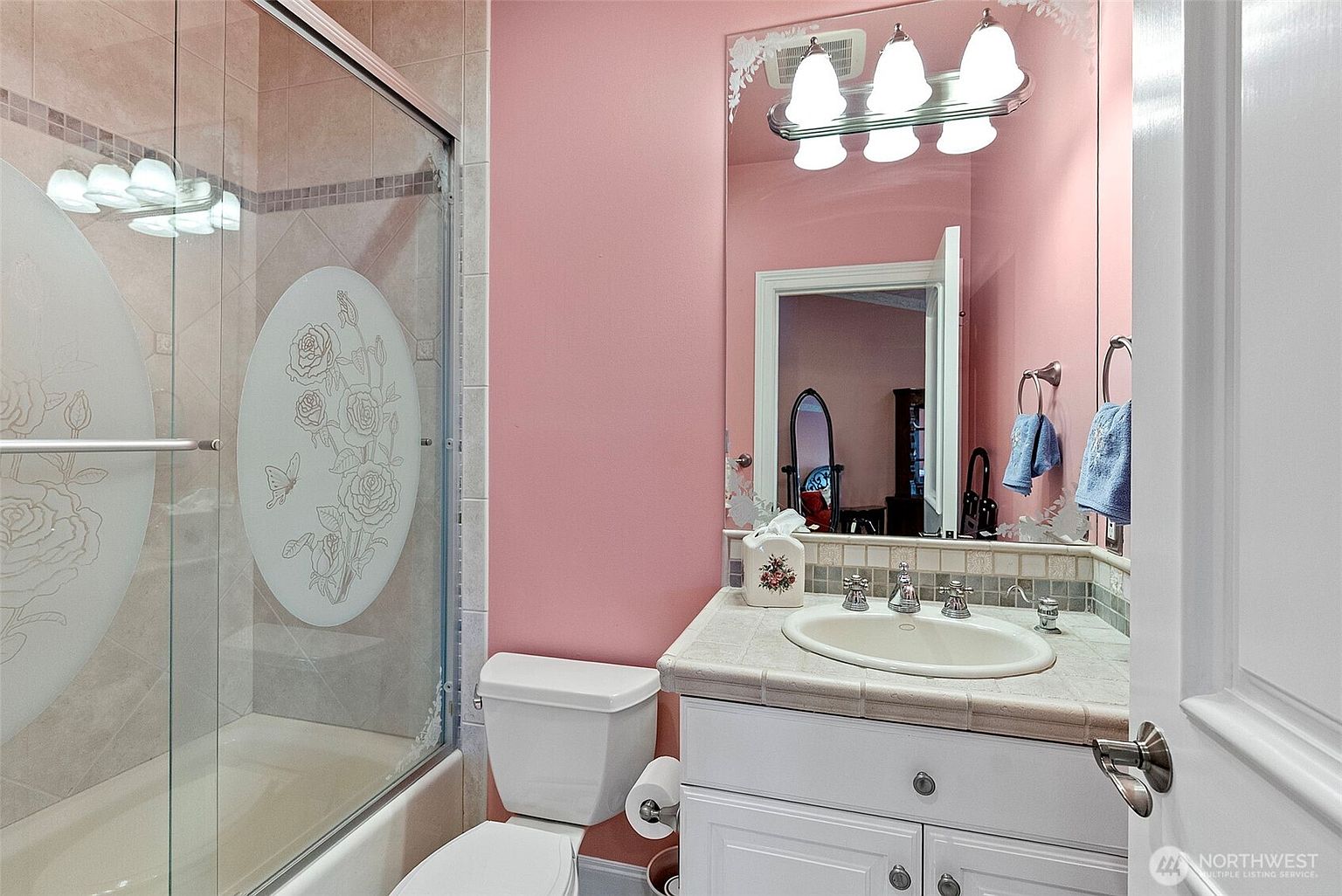 This bathroom features pink walls and a white vanity with a light-colored stone countertop and an oval undermount sink. To the left, a glass-enclosed shower stall is decorated with an etched floral design on the glass panel. A large mirror above the vanity reflects the room, complemented by a three-light fixture and blue hand towels hanging on the wall.