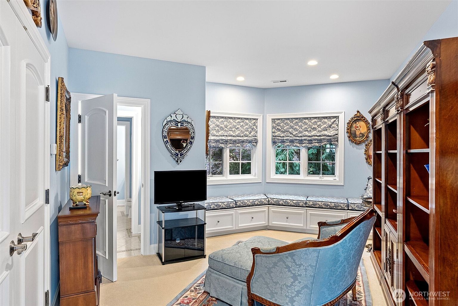 This cozy, light-filled room features soft blue walls and a built-in window seat with patterned cushions nestled into a bay window area. The space is furnished with a classic chaise lounge, a dark wood bookshelf, and a television on a glass stand, creating a comfortable reading or relaxation nook. A doorway leads into an adjacent bathroom, and decorative framed art adorns the walls, contributing to a traditional and inviting aesthetic.