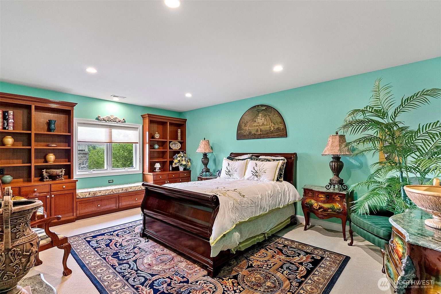This spacious primary bedroom features vibrant teal walls and a traditional dark wood sleigh bed as the central focal point. The room is appointed with built-in wooden shelving units flanking a window seat, a large ornate area rug, and two bedside tables with matching lamps. A large potted plant adds a touch of greenery to the corner, creating a warm and classic atmosphere.