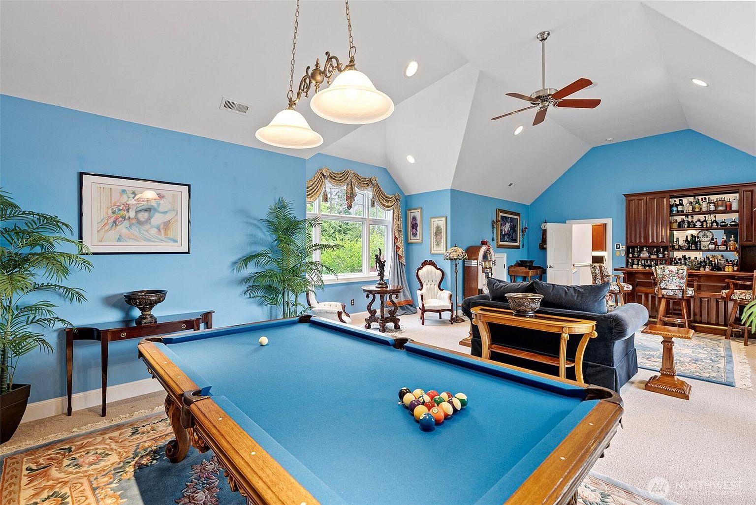 This spacious game room features a prominent blue pool table as the central focal point, set against vibrant blue walls and a vaulted ceiling. The room includes a fully equipped wooden bar area in the background, a comfortable seating area with a dark sofa, and a decorative jukebox, creating an ideal space for entertainment. Large windows with ornate valances allow natural light to fill the room, which is accented by a patterned area rug and classic pendant lighting.