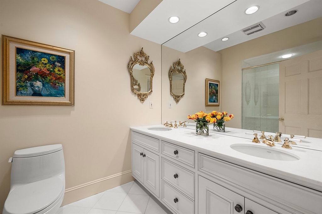 This is a well-lit bathroom featuring a double vanity with white cabinetry and a white countertop. Ornate gold-framed mirrors hang above each sink, and a framed painting adds a touch of color to the wall. A toilet is visible to the left, and the overall impression is clean and classic.