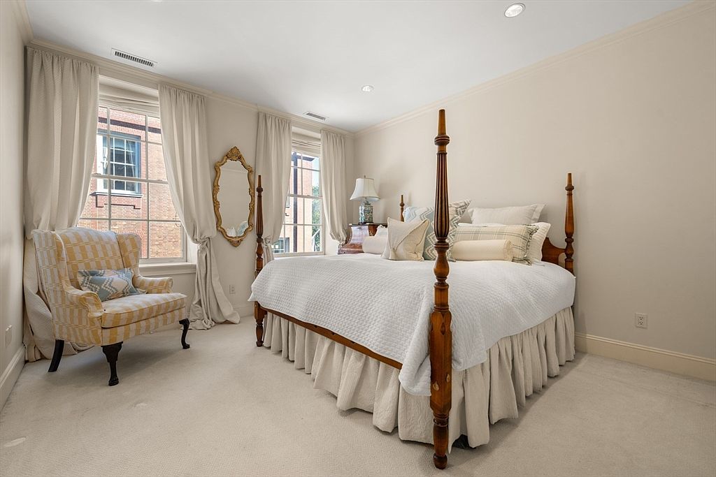This is a well-lit bedroom featuring a four-poster bed with white bedding and decorative pillows. The room has neutral-colored walls, carpeted flooring, and two windows with light-colored curtains. An armchair sits near one of the windows, and a mirror is positioned next to the bed, creating a cozy and inviting atmosphere.