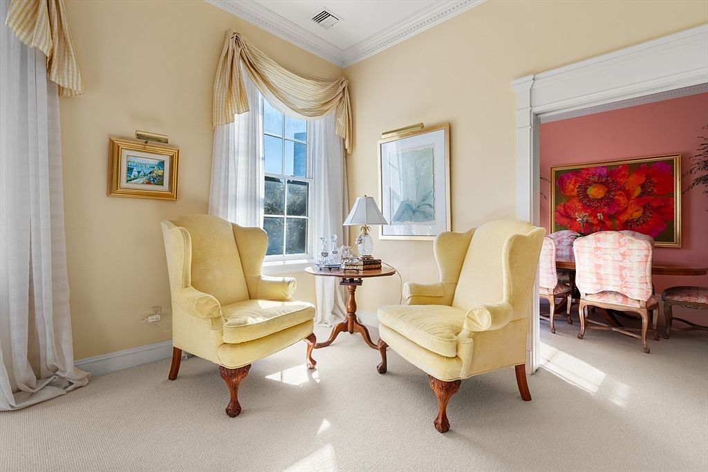 This interior shot showcases a cozy living room area with two yellow armchairs flanking a small round table. Natural light streams in through a window adorned with elegant drapery, highlighting the room's warm color palette and traditional style. A glimpse into an adjacent room reveals a dining area with floral artwork, adding depth and interest to the composition.
