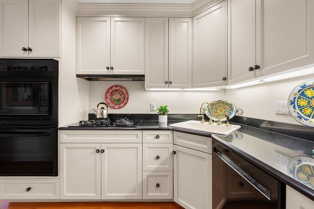 This is a well-lit kitchen featuring white cabinets with black hardware, black countertops, and stainless steel appliances. The kitchen includes a built-in oven and microwave, a gas stovetop, and a sink with a gold faucet. Decorative plates add a touch of personality to the space.