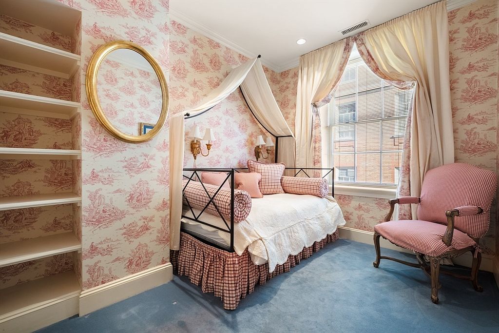 This charming guest bedroom features a daybed with a canopy, adorned with patterned pillows and a ruffled bed skirt. The walls are covered in a classic toile wallpaper, complemented by a gold-framed oval mirror and built-in shelving. A window with elegant drapes provides natural light, and a patterned armchair adds a cozy touch, all set against a blue carpet.