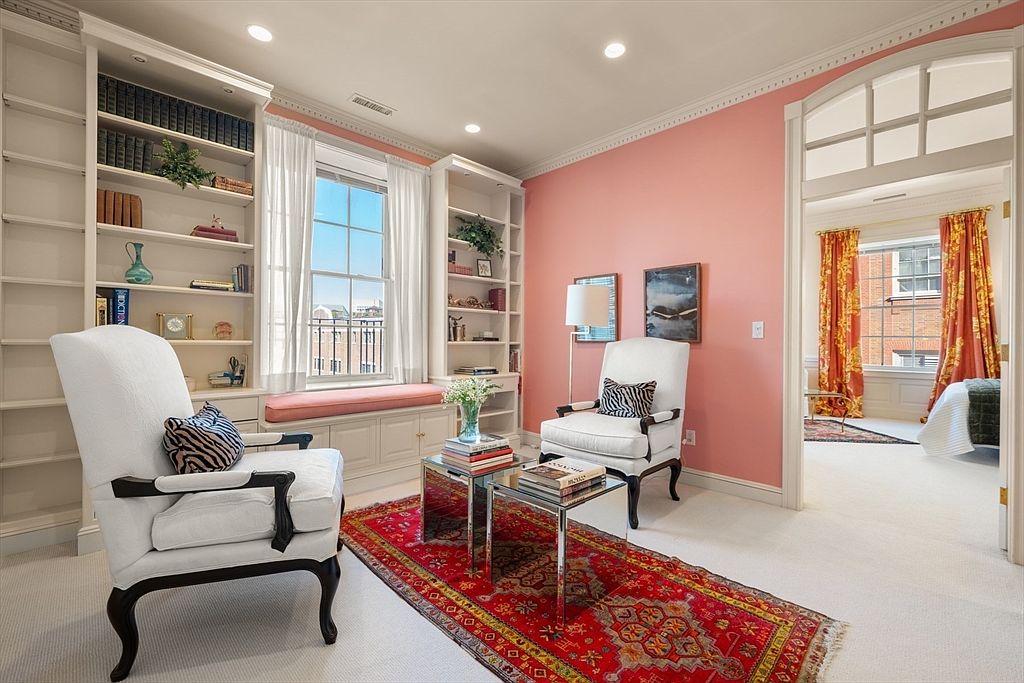 This interior shot showcases a cozy office or study area with built-in bookshelves flanking a window seat. Two elegant white armchairs face each other across a red patterned rug, with mirrored side tables in between. The room features a soft pink wall color, adding warmth and character to the space, while an open doorway leads to another room, creating a sense of flow and connectivity.