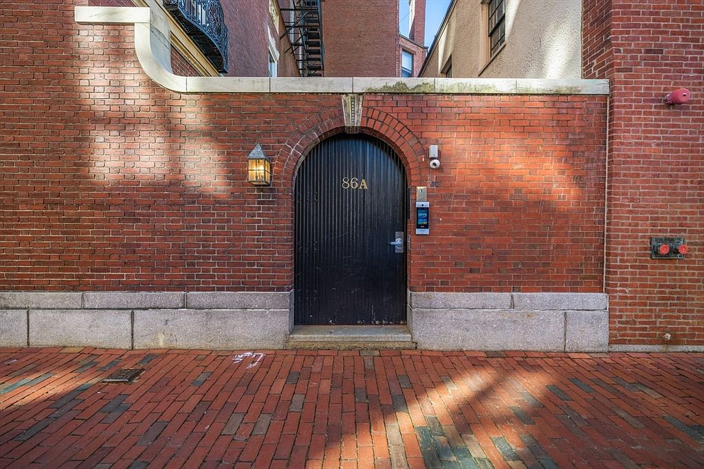 This image showcases an exterior entryway featuring a dark, arched door labeled '86A' set within a red brick facade. The entrance is flanked by a stone base and accented by a vintage-style lantern. The brick-paved ground and the surrounding brick walls create a charming, urban aesthetic, suggesting a well-maintained and secure property.