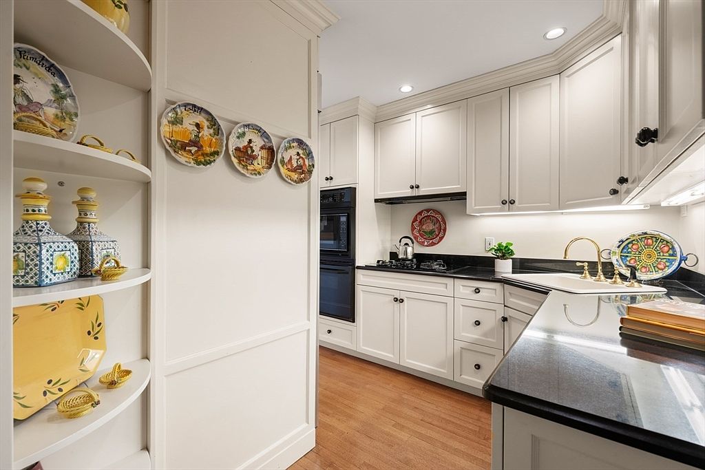 This is a well-lit kitchen featuring white cabinetry, black countertops, and hardwood flooring. Decorative plates adorn the walls, adding a touch of personality to the space. The kitchen is equipped with modern appliances, including a built-in oven, and a gold faucet adds a touch of elegance.
