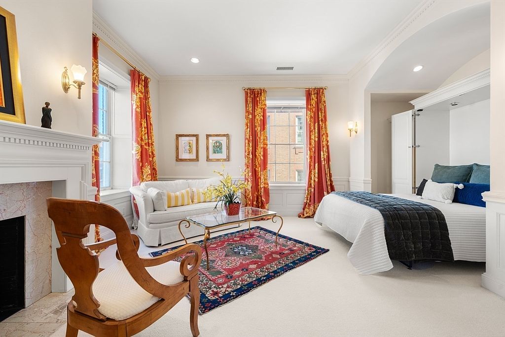 This is a well-appointed guest bedroom featuring a Murphy bed, a comfortable sofa, and a decorative fireplace. The room is bright and airy, with natural light streaming through the windows, complemented by elegant curtains. A patterned rug adds warmth and character to the space, creating a welcoming atmosphere.