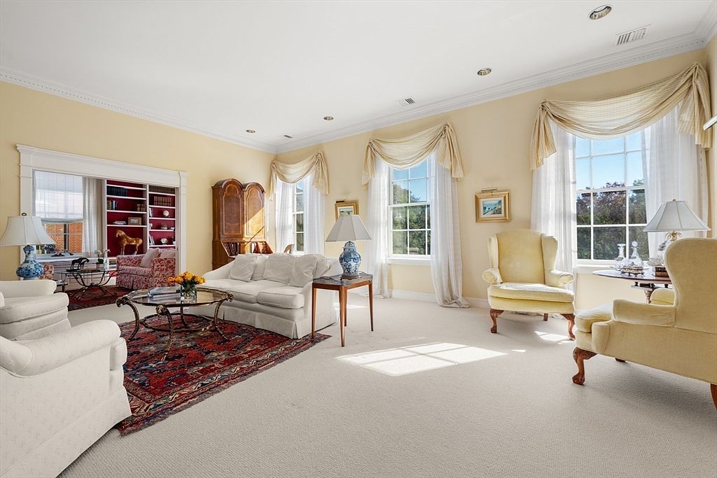 This is a bright and elegant living room featuring a neutral color palette with yellow walls and light beige carpeting. The room is furnished with a white sofa, two yellow armchairs, and a glass-topped coffee table, complemented by traditional-style lamps and window treatments. The space is well-lit with natural light streaming through the large windows, creating a warm and inviting atmosphere.
