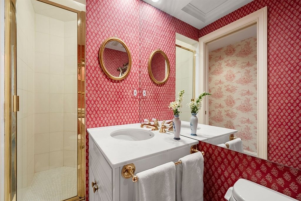 This is a guest bathroom featuring red patterned wallpaper, a white vanity with a marble countertop, and gold fixtures. The bathroom includes a shower with a gold frame and a separate toilet area, creating a luxurious and coordinated aesthetic. The oval mirrors and decorative vases add to the room's charm.