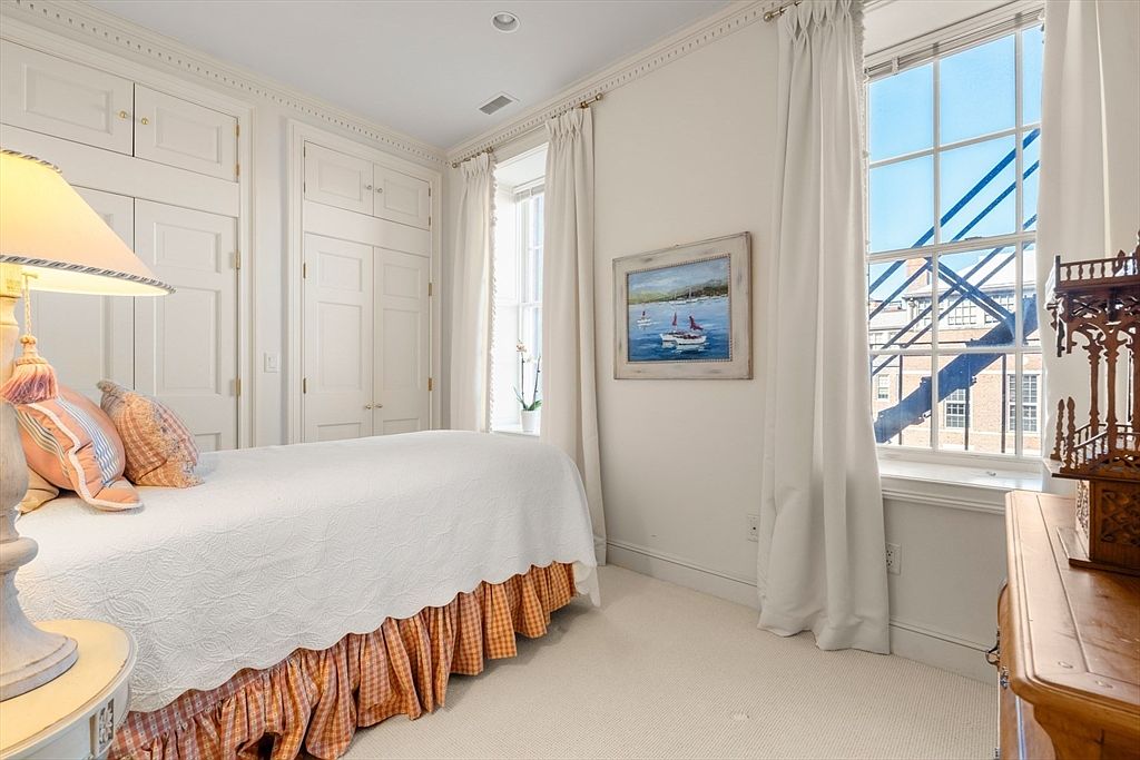 This is a cozy guest bedroom featuring built-in closets and a single bed with a white quilted coverlet and orange patterned bed skirt. A painting hangs on the wall, and natural light streams in through the window, which is framed by white curtains. The room has a classic and comfortable feel.