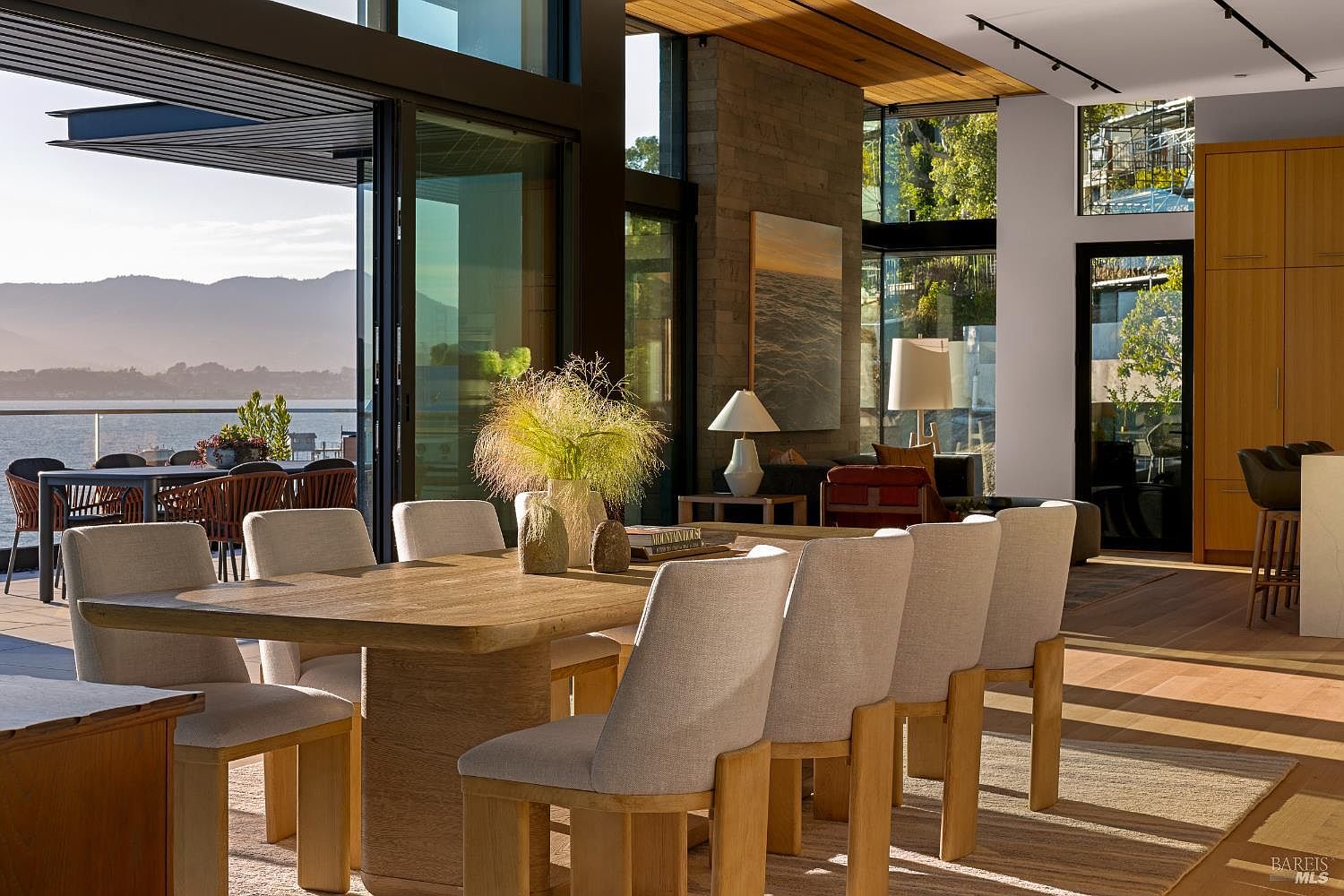 This sophisticated dining area features a large light wood table surrounded by upholstered chairs, set against a backdrop of expansive glass walls that frame a stunning waterfront view. The open-concept space flows seamlessly into a living area with a stone-clad chimney breast and warm wood ceilings, creating a bright and airy atmosphere. The perspective captures the transition between the elegant indoor dining space and the outdoor deck, emphasizing the home's connection to the natural landscape.